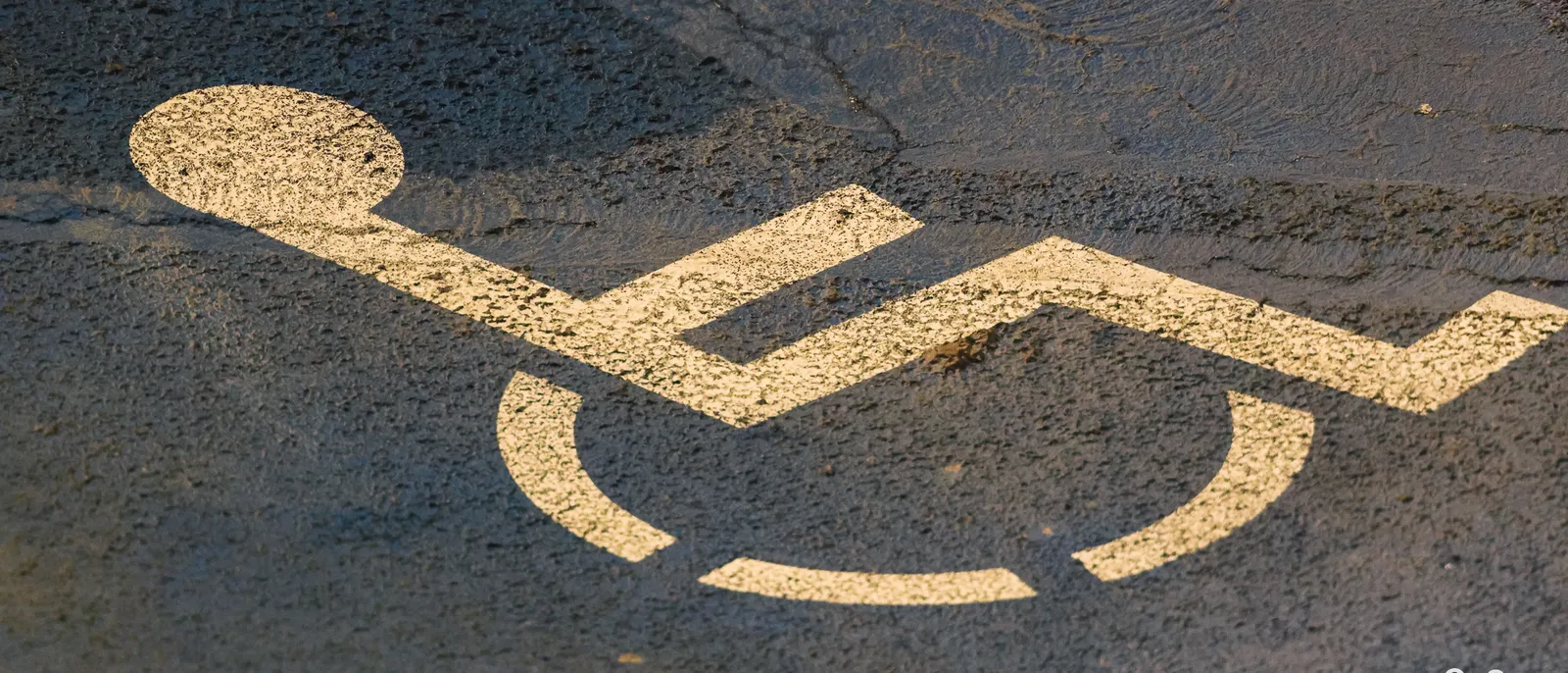 Pictogramme fauteuil roulant peint en blanc sur l’asphalte d’un parking, éclairé par une lumière rasante.