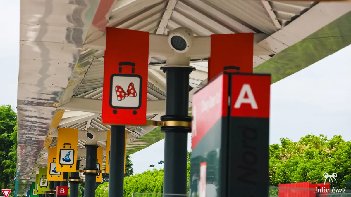 Covered bays of the north coach station at Disneyland Paris, with hotel signs (Minnie bow, wizard hat) and the bay A post in front