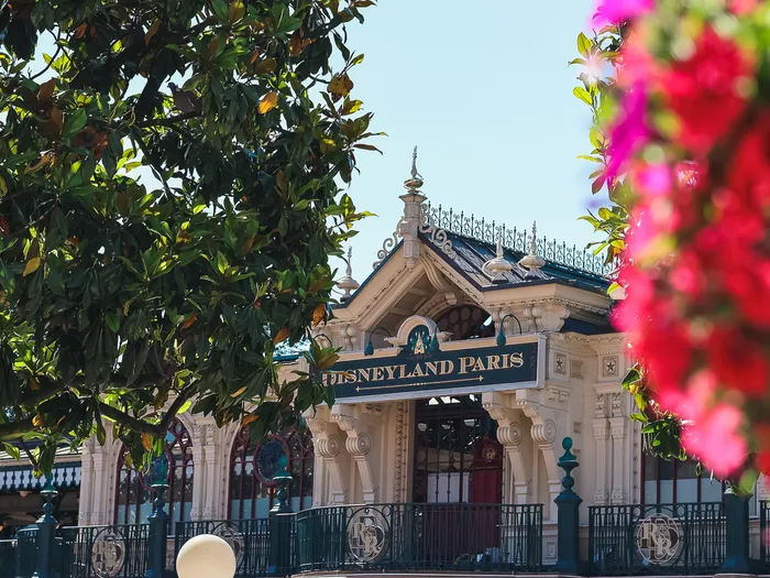 Façade blanche de la Main Street Station et son enseigne Disneyland Paris, encadrées par un arbre et des fleurs roses.