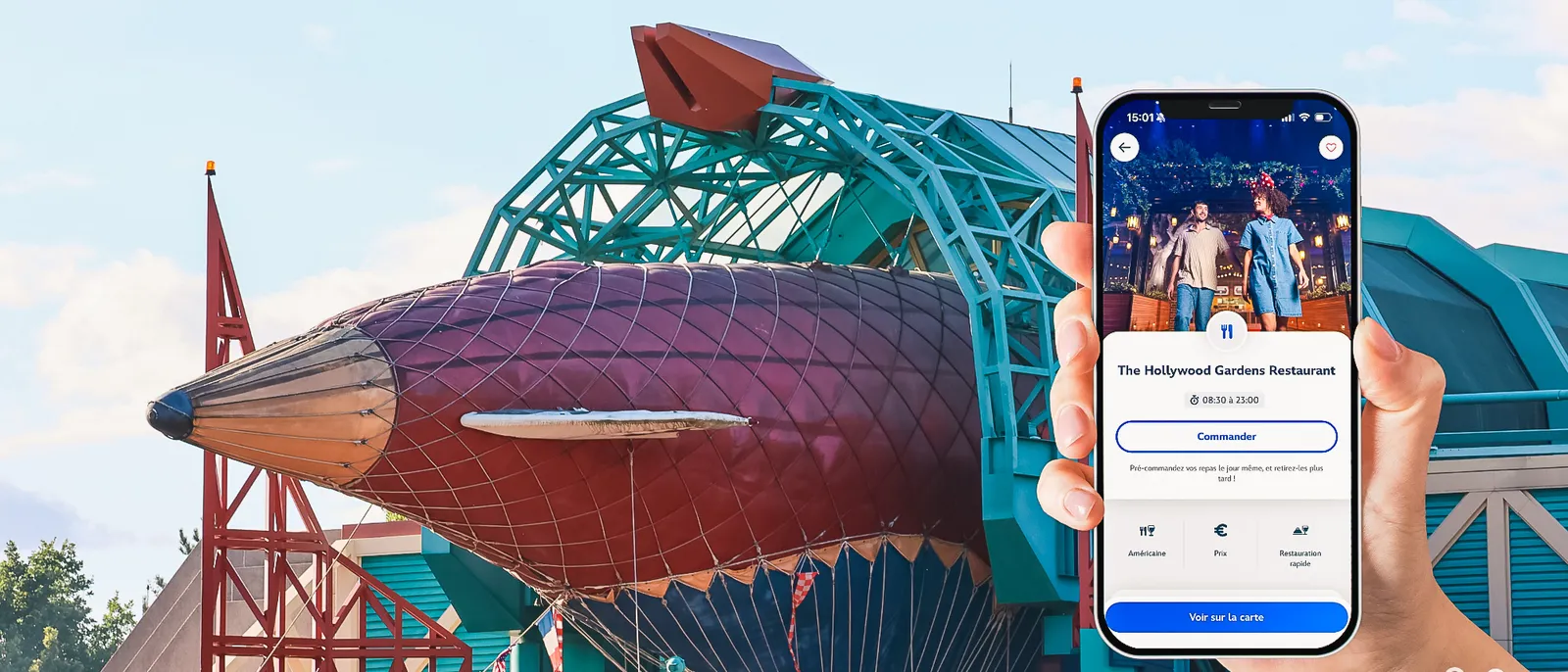 Main tenant un smartphone, écran de commande Click and Collect de l’app Disneyland Paris devant un bâtiment du parc