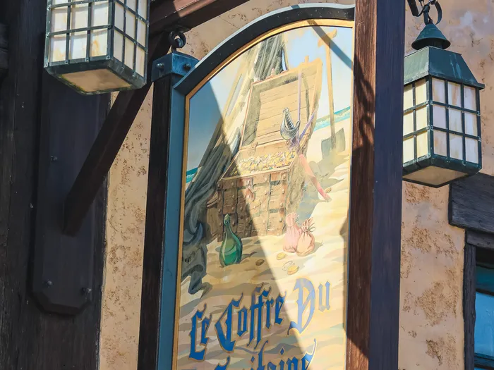 Painted hanging sign of the Le Coffre du Capitaine shop at Disneyland Paris, framed by two lanterns and illustrated with a pirate treasure scene