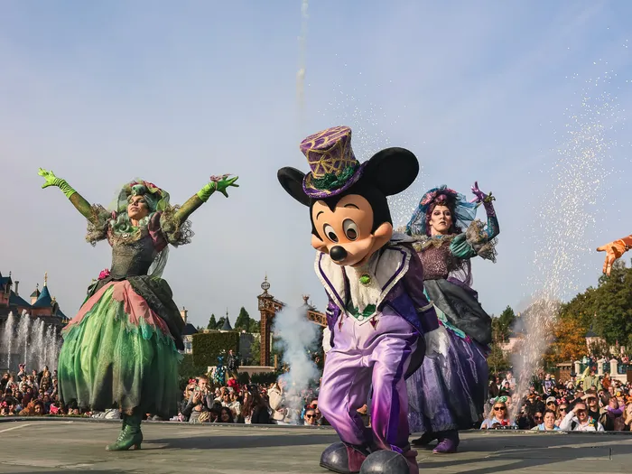 Mickey en costume violet et haut-de-forme danse avec deux artistes sur scène devant le château, spectacle Halloween du Parc Disneyland.