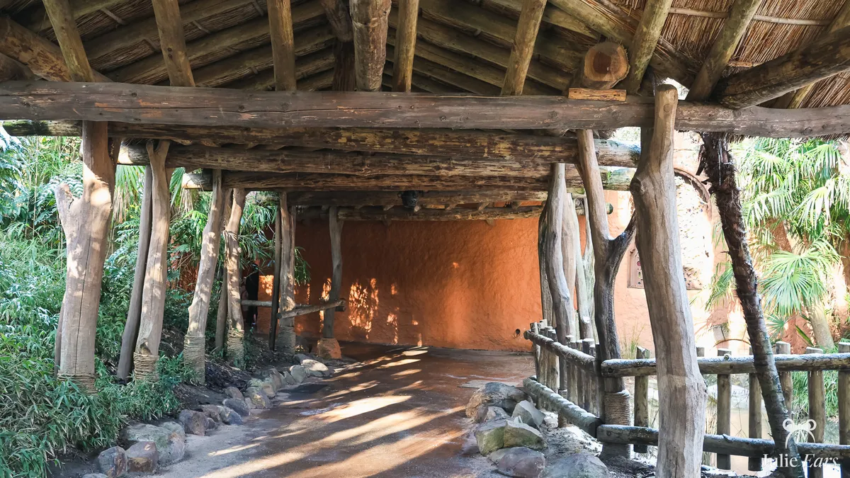 Passage couvert d’un toit de chaume porté par des troncs bruts, côté Adventureland à Disneyland Paris, avec un sol tacheté de soleil bordé de pierres, un mur ocre orangé et une végétation tropicale.