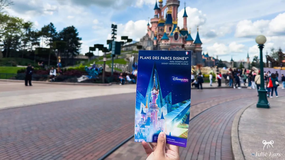 Brochure Plans des Parcs Disney tenue à la main devant le château du Parc Disneyland