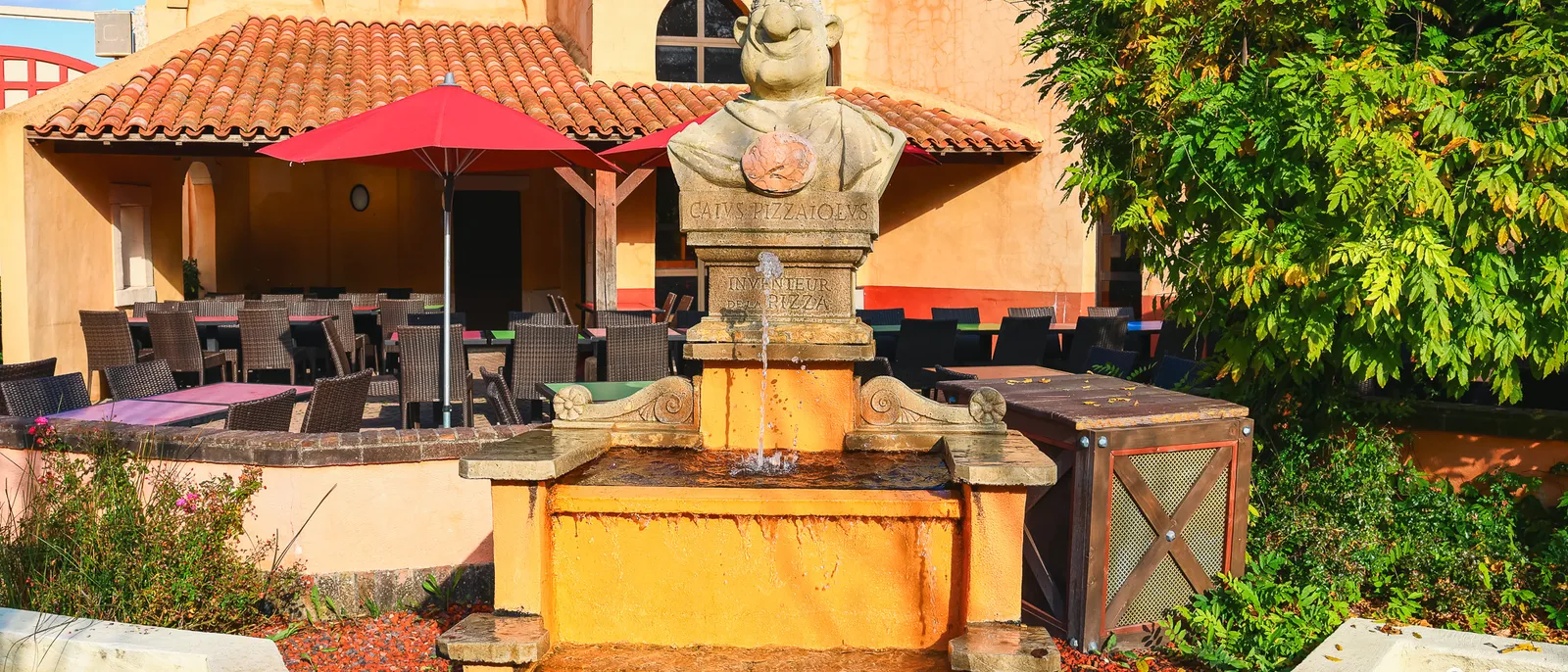 Fontaine surmontée d’un buste devant la terrasse du Caius, façade ocre à tuiles rondes et parasol rouge déployé