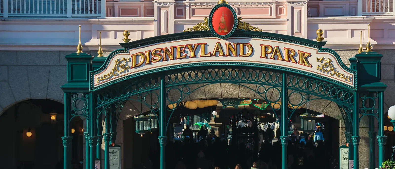 Arche en fer forgé vert portant l’inscription « Disneyland Park », visiteurs entrant sous le Disneyland Hotel rose au soleil.