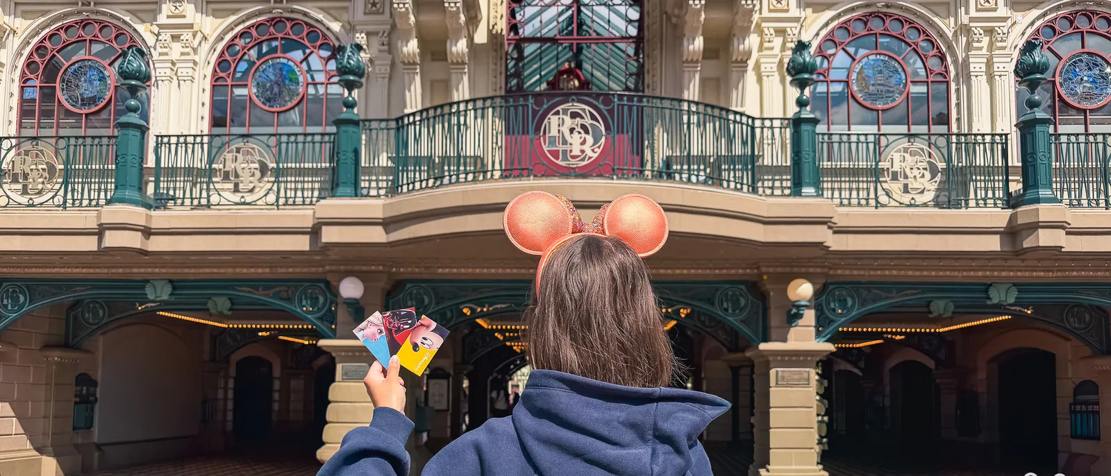 Visiteuse de dos avec oreilles Minnie brandissant ses billets devant la façade Disneyland Paris de la gare.