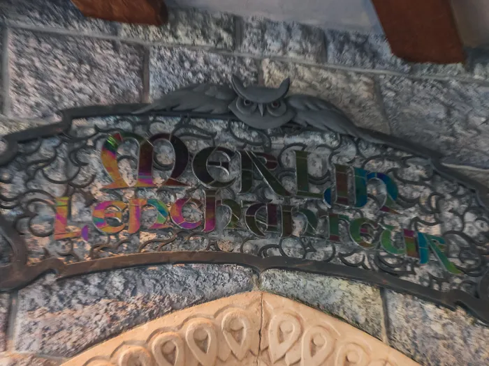 Wrought-iron sign with coloured Merlin l’Enchanteur lettering on the castle’s stone wall, at Disneyland Paris.