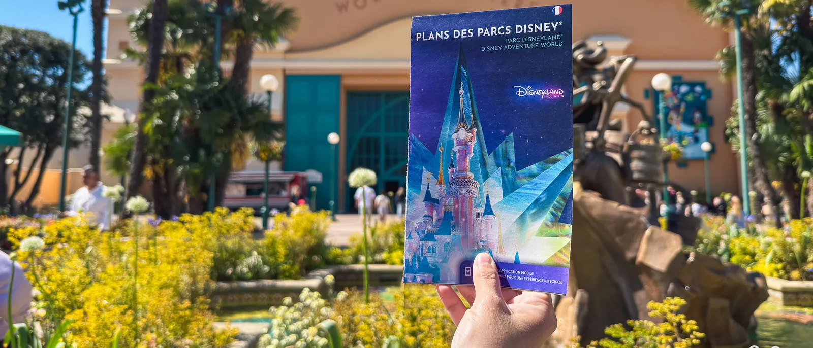 main tenant le plan papier des deux parcs devant l’entrée du Parc Disney Adventure World, parterres fleuris.