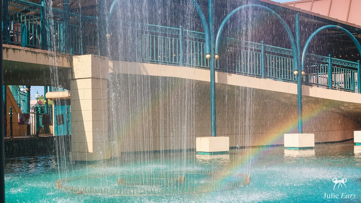 Water jets making a rainbow in the turquoise pool below the "it’s a small world" walkway, in Fantasyland at Disneyland Park, on a sunny day