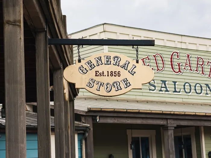 Oval "General Store, Est. 1856" sign hanging from a bracket in front of a wooden Western frontage.