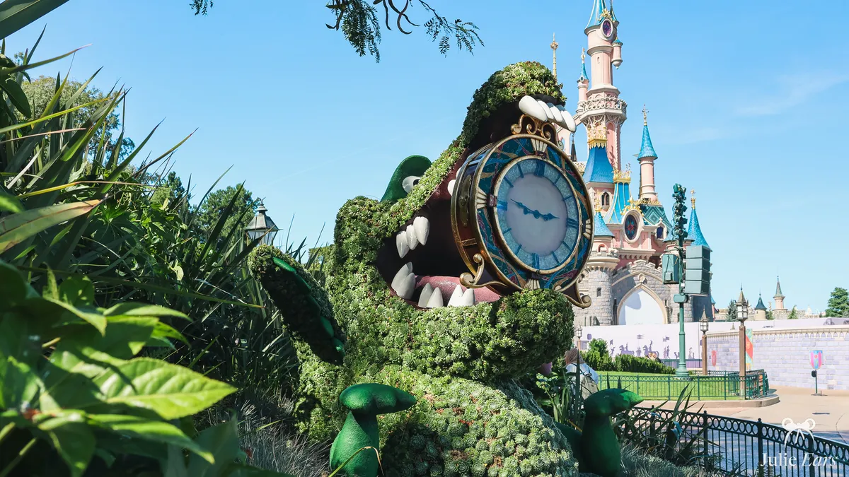 Topiary of Tick-Tock the crocodile from Peter Pan, a clock in his jaws, in front of Sleeping Beauty Castle under a blue sky, at Disneyland Paris