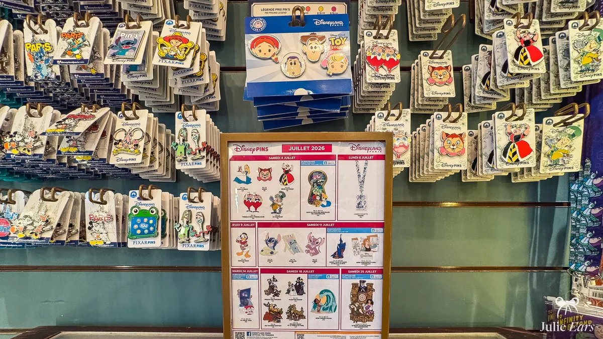 Disney Pins programme for July 2026 framed on a shop counter at Disneyland Paris, listing the releases by date, in front of a wall of pins