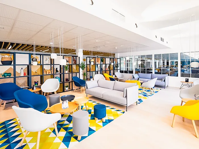 Salon commun du Staycity Aparthotel avec fauteuils bleus et jaunes, canapés gris et tapis géométrique coloré.