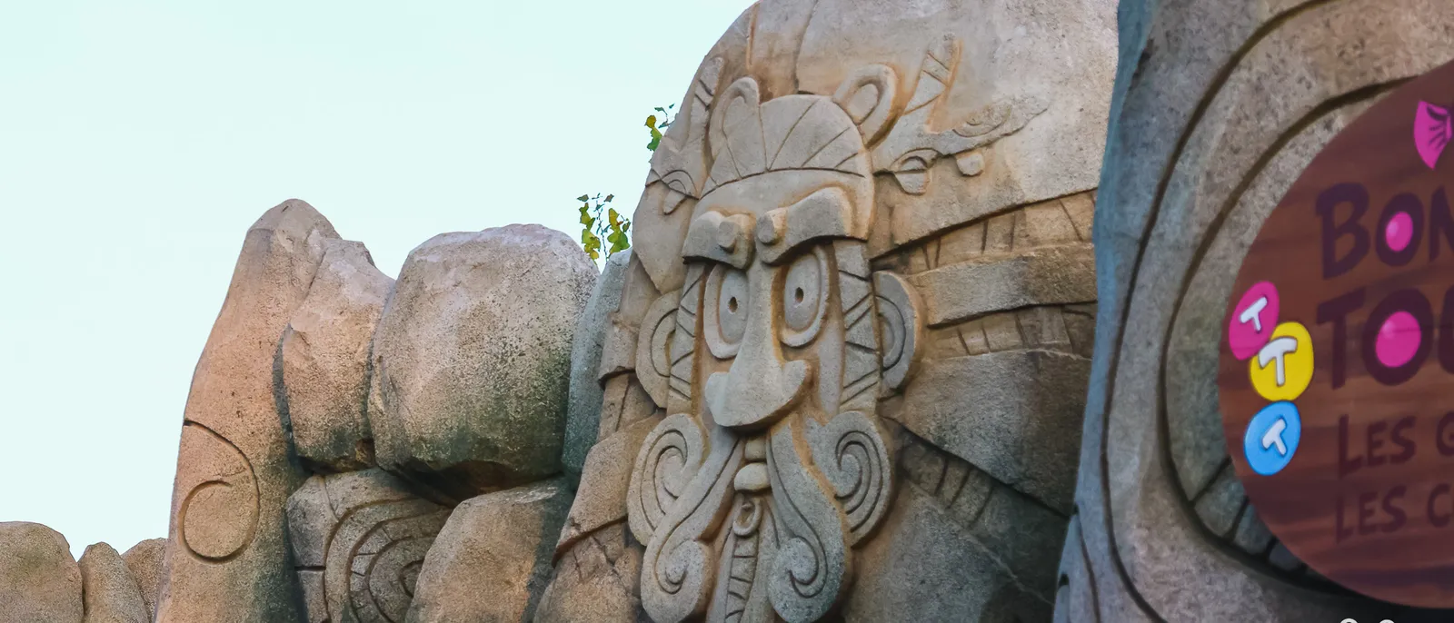 Visage barbu de Toutatis sculpté dans la roche à l'entrée du coaster, panneau rouge et pastilles colorées à droite
