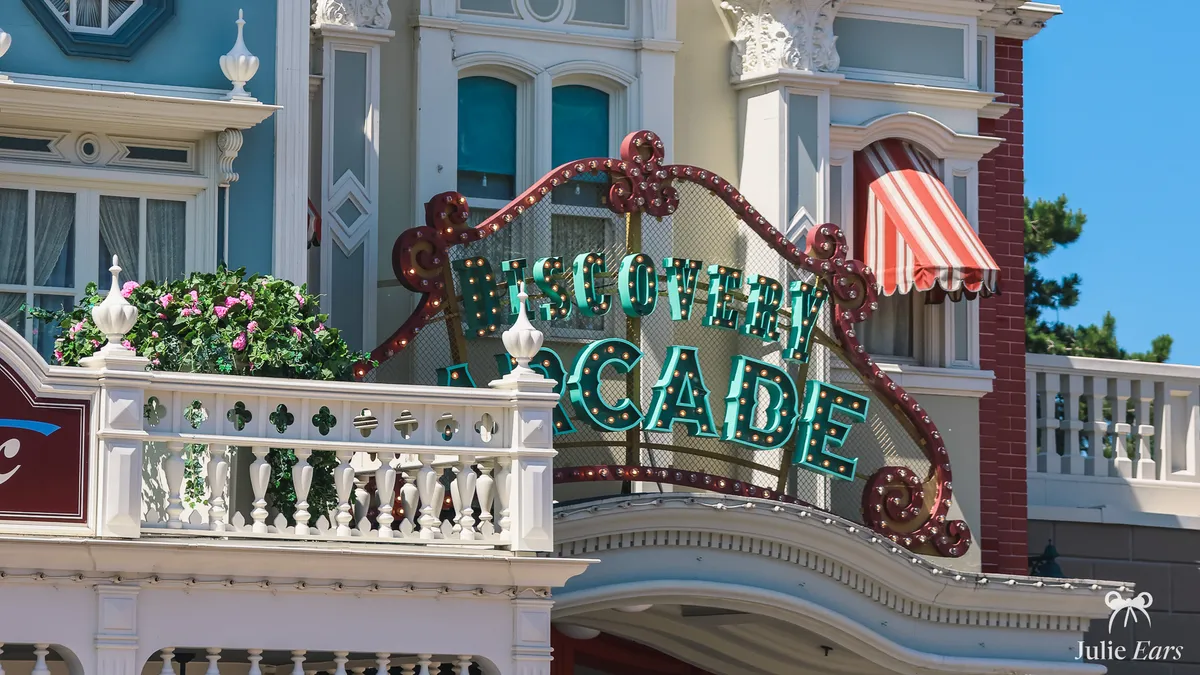 Enseigne "Discovery Arcade" aux lettres turquoise serties d’ampoules dans un cadre rouge à volutes, derrière un balcon blanc fleuri et entre des façades victoriennes de Main Street U.S.A. à Disneyland Paris.