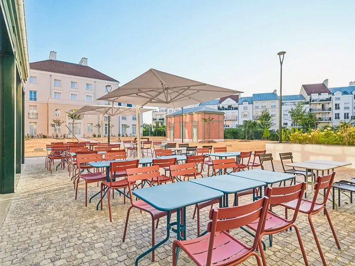 Terrasse extérieure de l’Appart Hôtel All Suites Val d’Europe, tables et chaises colorées sous un parasol, dans le quartier de Val d’Europe.