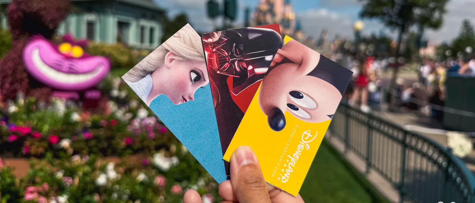 Trois billets Disneyland Paris tenus devant le château, avec la topiaire du Chat du Cheshire et les massifs fleuris.