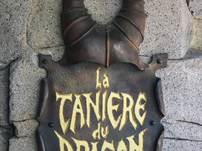 Enseigne dorée « La Tanière du Dragon » surmontée de cornes de Maléfique, fixée sur la roche du château.