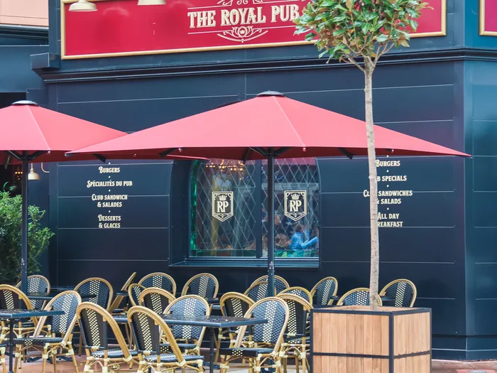 Terrasse du Royal Pub au Disney Village avec chaises en rotin, parasols rouges et façade noire portant l’enseigne dorée.