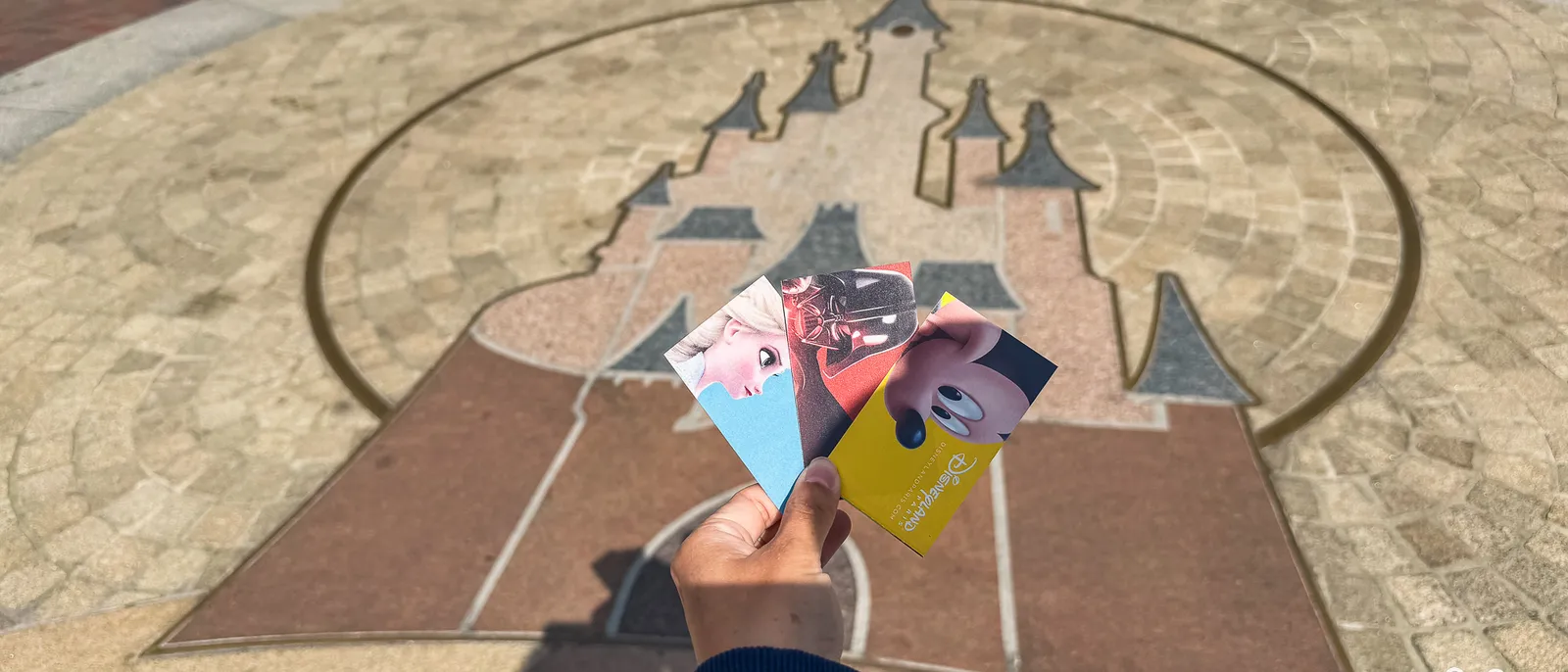 Main tenant trois billets Disneyland Paris au-dessus de la mosaïque au sol dessinant le château.