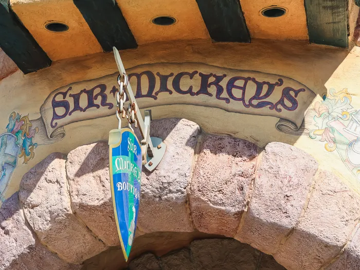Enseigne en bois de la boutique Sir Mickey’s sous une arche de pierre claire, à Fantasyland.