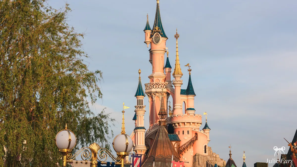 Pink towers and blue-green spires of Sleeping Beauty Castle at Disneyland Paris in golden light, with a willow and a golden lamppost in front