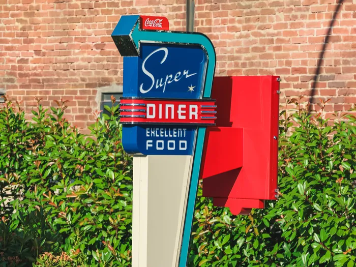 Panneau rétro bleu et rouge « Super Diner, Excellent Food » devant un mur de briques et des buissons