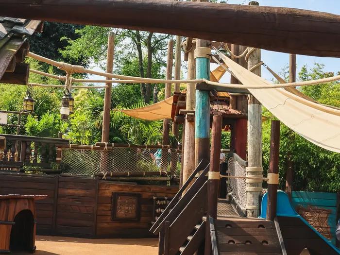 Aire de jeux Plage des Pirates avec mâts de bois, voiles tendues et toboggan sur sol souple, en plein soleil