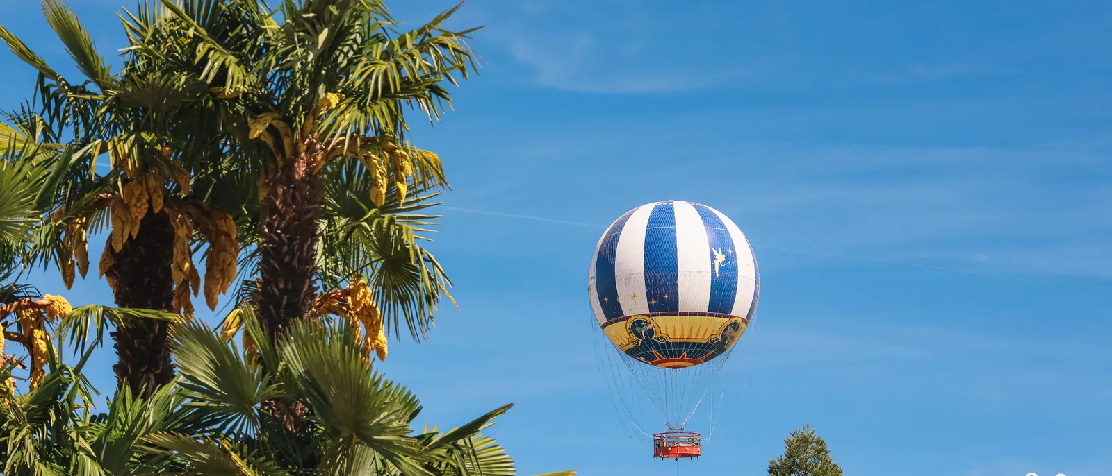 Ballon captif blanc et bleu du PanoraMagique flottant dans un ciel dégagé, à côté de palmiers du Disney Village.