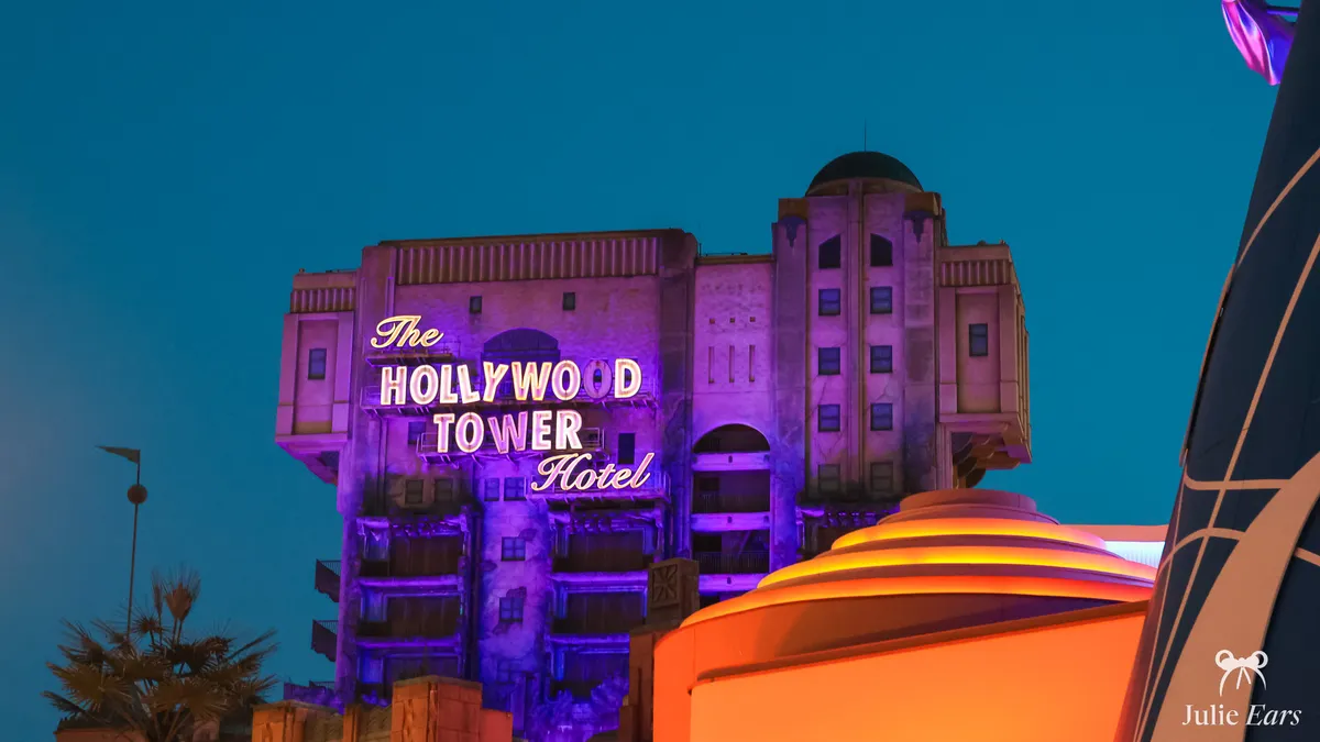 The Hollywood Tower Hotel lit up in purple at blue hour, its sign switched on, behind a rounded roof lit in orange at Disney Adventure World