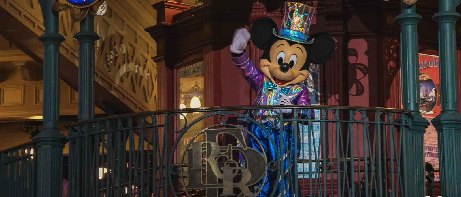 Mickey en costume à paillettes et haut-de-forme salue de la main depuis un balcon ouvragé, Disneyland Paris.