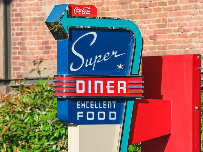 Enseigne néon rétro bleu et rouge « Super Diner Excellent Food » devant un mur de briques, Parc Disney Adventure World.