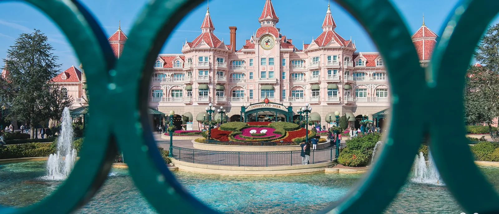 The Disneyland Hotel seen through a railing with green rings, with the fountains and the Mickey flower bed in front of the park entrance