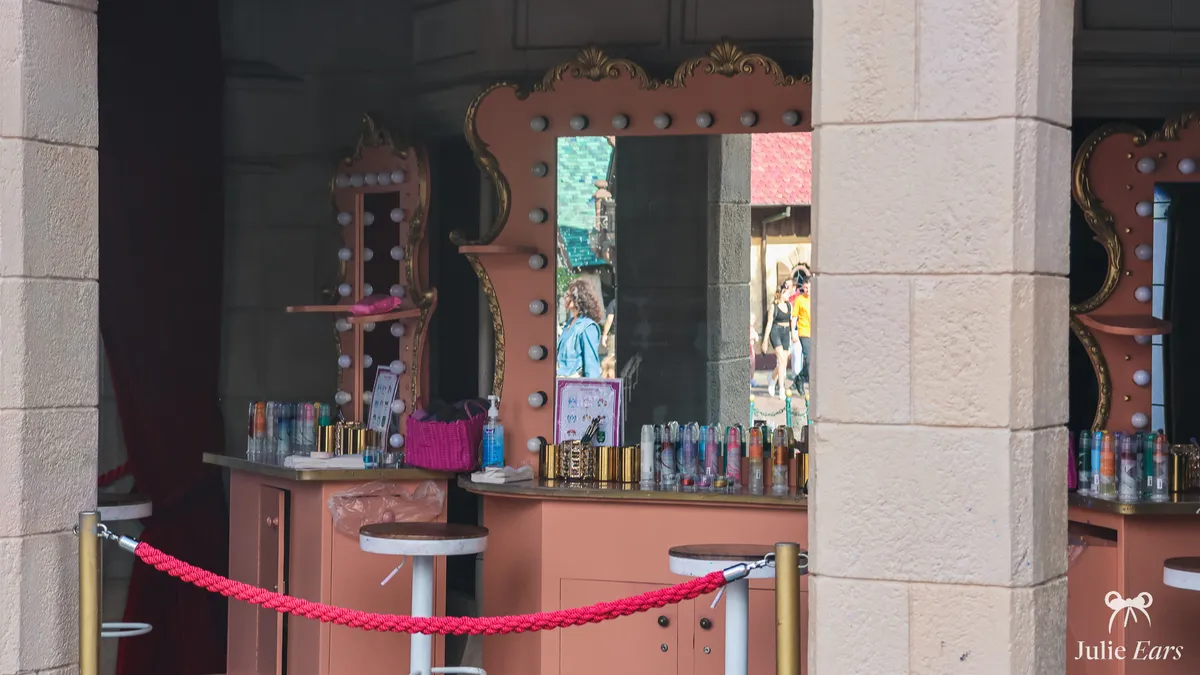 Stand de maquillage en extérieur à Fantasyland, avec ses miroirs à ampoules, ses tabourets et les produits alignés sur le comptoir.