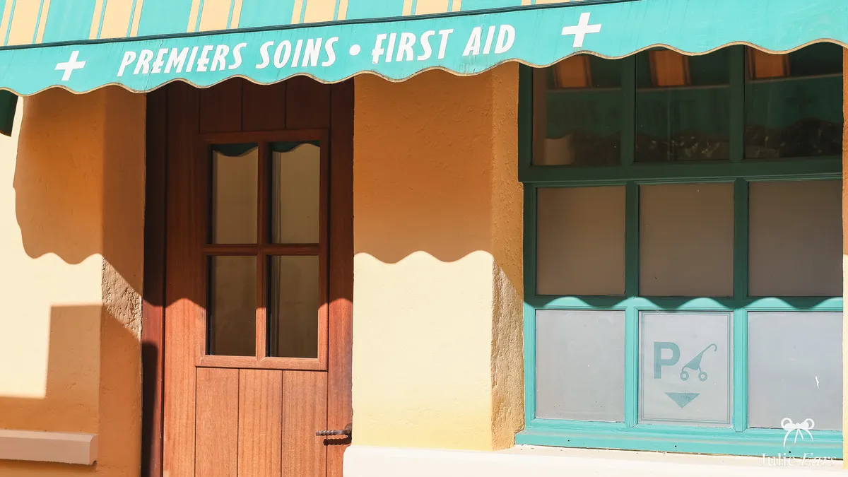Turquoise awning reading "PREMIERS SOINS • FIRST AID" at Disney Adventure World, above a wooden door and a window with the pushchair parking pictogram