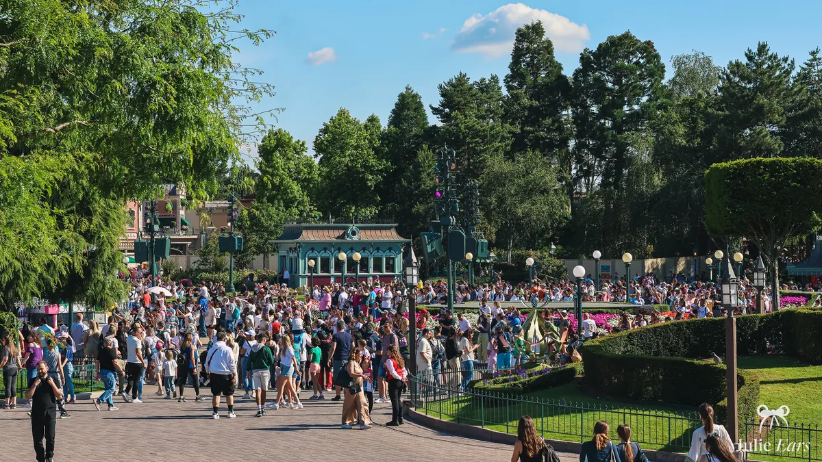 Dense crowd on Central Plaza in Disneyland Park, Disneyland Paris, between flower beds, trimmed hedges and a green pavilion on a sunny day