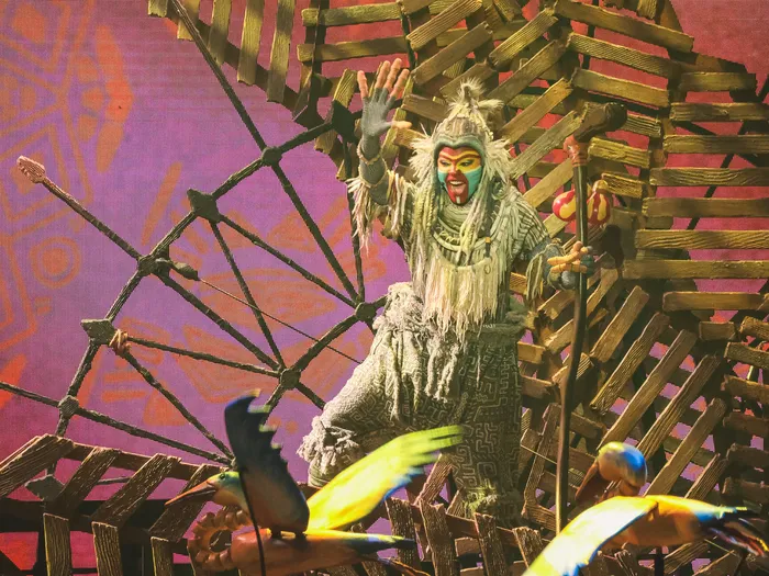 Comédien costumé en chaman bras levés sur une structure en bois avec des oiseaux, tableau Roi Lion du spectacle Mickey et le Magicien