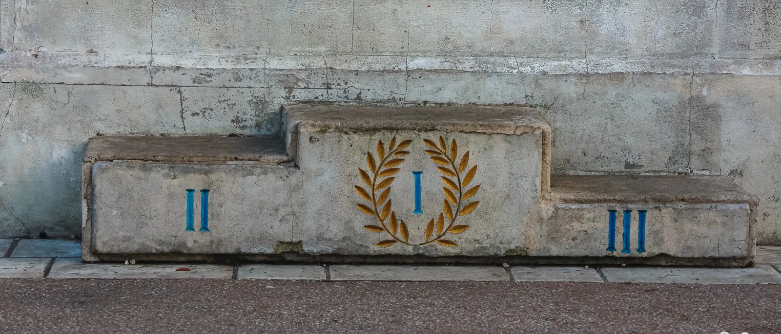 Podium en béton du Parc Astérix, marches I, II et III en chiffres romains bleus et couronne de laurier dorée peinte