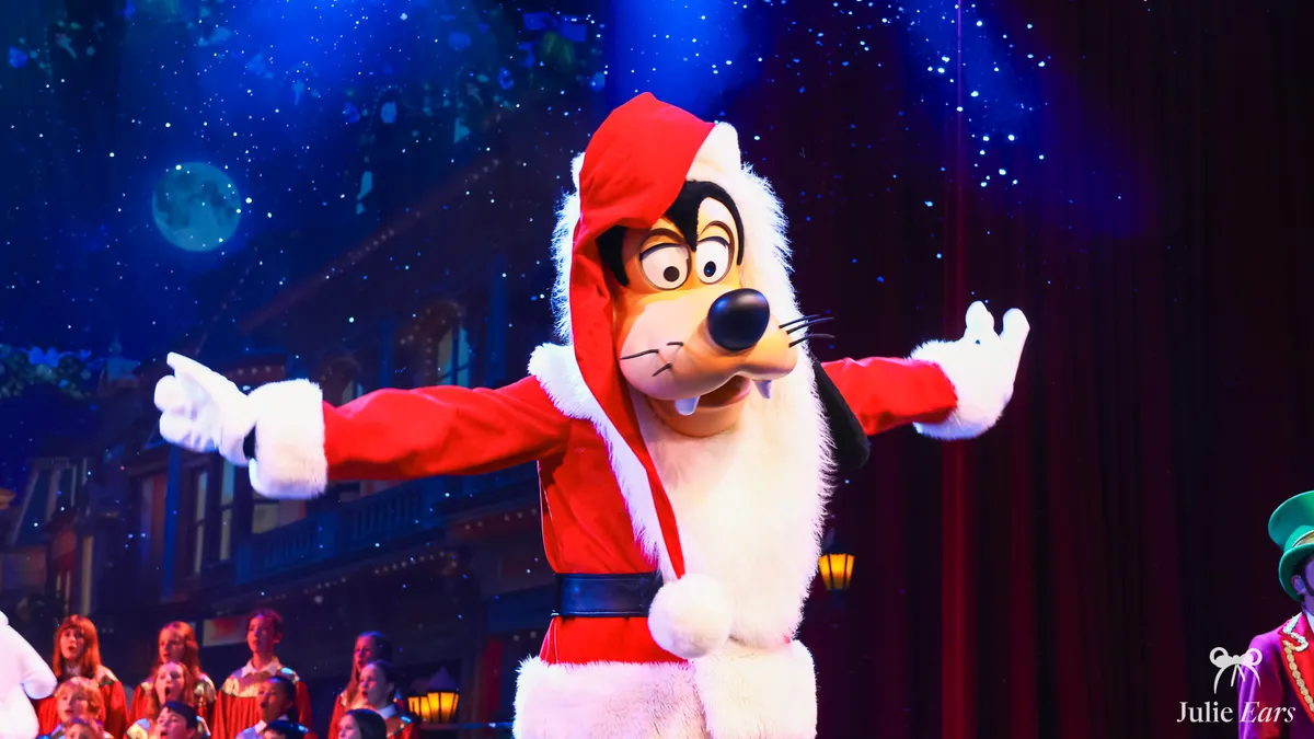 Dingo déguisé en Père Noël, bras écartés sur scène pendant le spectacle Chantons Noël à Disneyland Paris