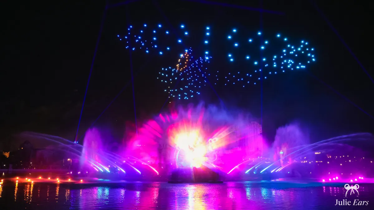 Disney Cascade of Lights at night: drones draw glowing blue shapes in the sky above the pink and purple fountains of the Disney Adventure World lake