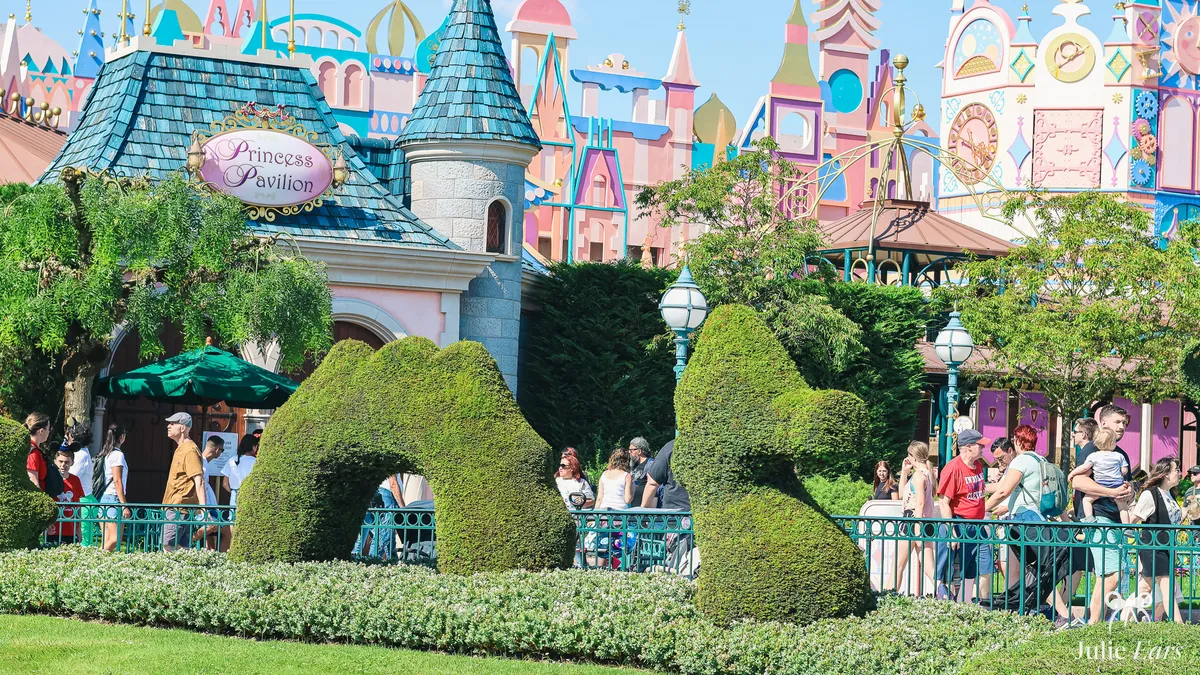 Princess Pavilion and its sign in Fantasyland at Disneyland Paris, behind topiaries, in front of the colourful "it’s a small world" facade