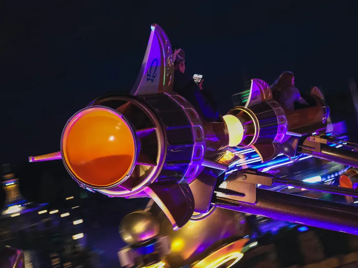 Gros plan sur une nacelle fusée d’Orbitron au hublot orange lumineux, saisie en mouvement dans la nuit à Discoveryland.