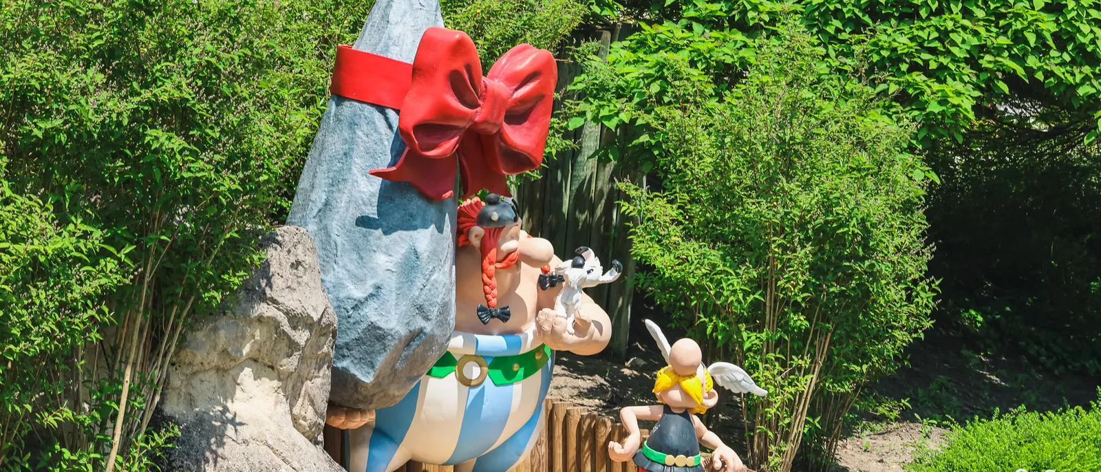 Statues d’Obélix portant un menhir noué d’un gros nœud rouge et d’Astérix casqué, devant la végétation