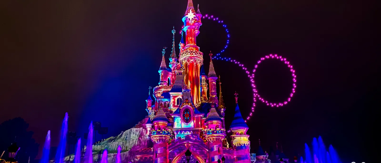 Château illuminé de rose et bleu la nuit, jets d’eau et chiffre "30" lumineux dans le ciel de Disneyland Paris.