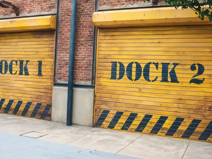 Deux rideaux métalliques jaunes marqués Dock 1 et Dock 2 sur un bâtiment de brique, Marvel Avengers Campus