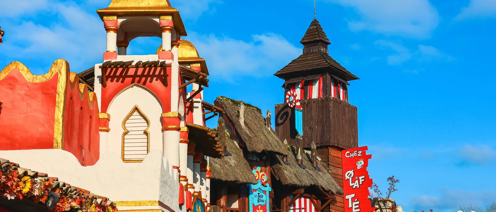 Façades colorées de la Via Antiqua : tour à dôme doré, toits de chaume et enseignes Touristix et Chez Olaf Tepaf sous un ciel bleu