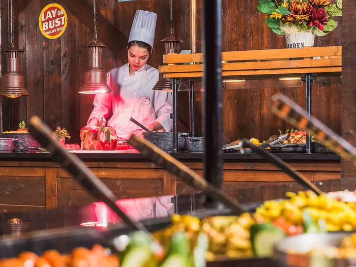 Cuisinière découpant une viande sous les lampes chauffantes du buffet rustique de La Grange, plats en premier plan.