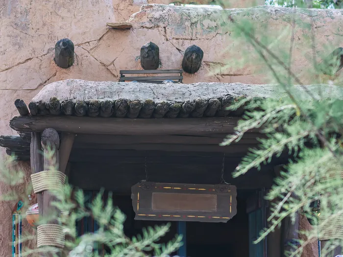 Adobe facade of Pueblo Trading Post in Frontierland, with jutting beams, a log awning and a wooden sign, seen through light foliage