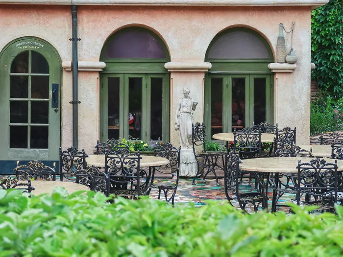 Terrasse de la Pizzeria Bella Notte avec tables et chaises en fer forgé autour d’une statue, devant les portes vertes du restaurant.